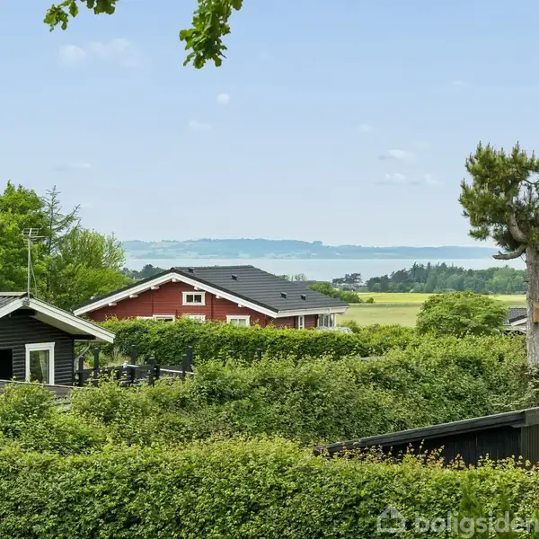 Flere huse omgivet af frodige buske og træer. I baggrunden ses en udsigt over en sø eller fjord med skovklædte bakker og blå himmel.