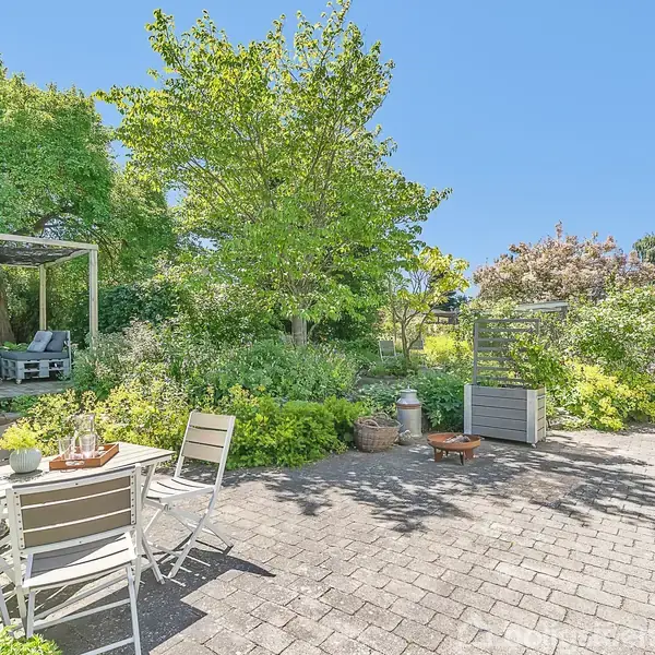 Et hvidt cafésæt står på en terrasse med fliser; omgivet af frodig have med træer og buske. I baggrunden ses en sofa under et pergola-tag.
