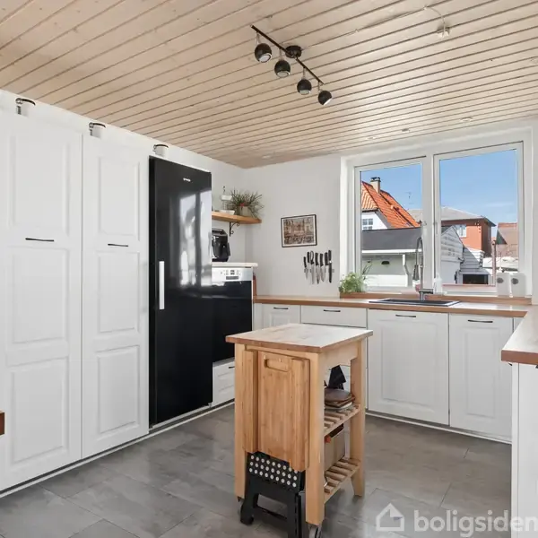 Et moderne køkken med hvide skabe og sorte apparater inkluderer en central træø. Et stort vindue giver udsigt til røde tage udenfor. Loftet har træbjælker og spotlys.