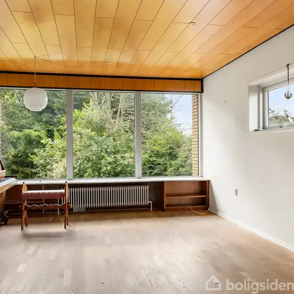 En fritstående brændeovn står i hjørnet af en stue med lys trægulv og hvidmalede vægge. Et lille vindue giver dagslys, og en lysekrone hænger fra loftet.