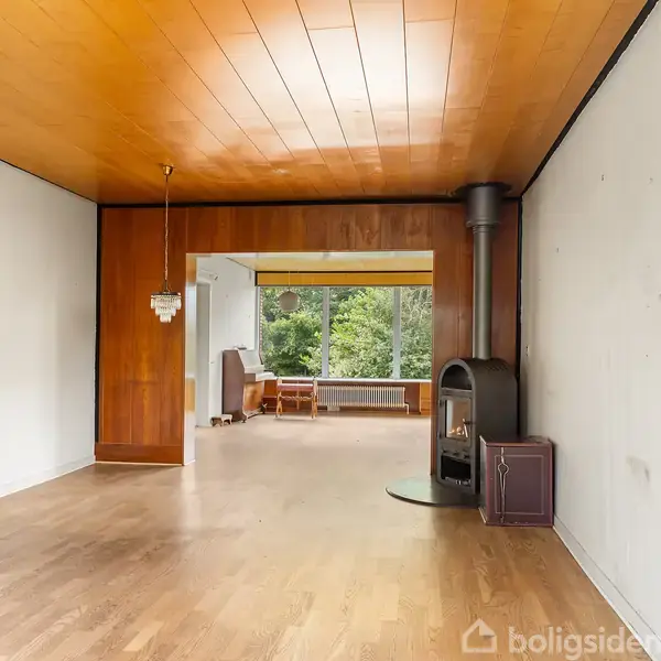 En brændeovn står i et rum med trægulv og træloft. I baggrunden ses store vinduer med udsigt til grønne træer og en lampe hænger fra loftet.