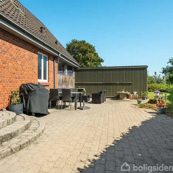 Rød murstensbygning med en terrasse, der har havemøbler og en grill. Terrassen er omgivet af en grøn have med planter og en høj, grøn bygning i baggrunden.