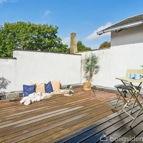 En træterrasse med to stole og et lille bord står foran en hvid mur. Der er puder og planter, mens grønne trætoppe ses bag muren, under en blå himmel.