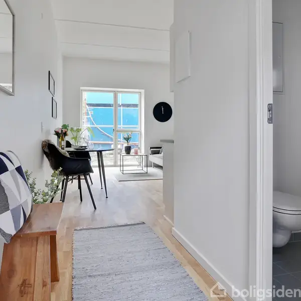 Træbænk med pude i en lys gang fører til en stue med spisebord og stole foran store vinduer. Til højre ses en delvist åben dør til et badeværelse.