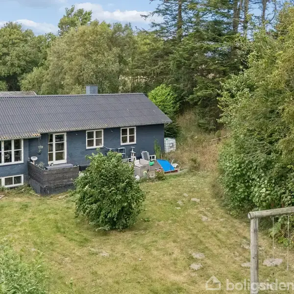 Et blåt hus står omgivet af grønne træer og buske i en frodig have med en græsplæne. En blå legeting ses ved siden af terrassen.