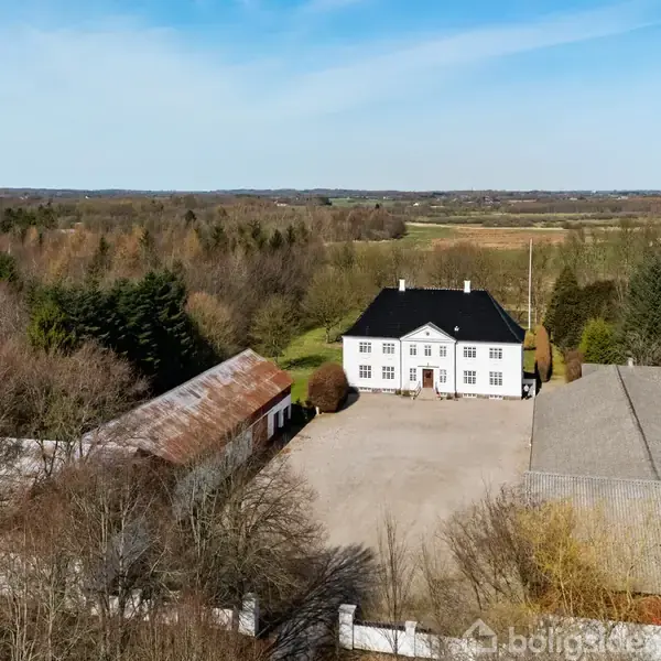 En stor hvid bygning med sort tag står stille omgivet af skove og marker. Til venstre er en rustik bygning og til højre en grå bygning.