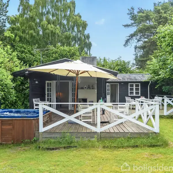 Et sort sommerhus med hvidt rækværk står på en terrasse, omgivet af grønne træer. Der er en parasol over havemøbler og et surfbræt, samt en pool til venstre.