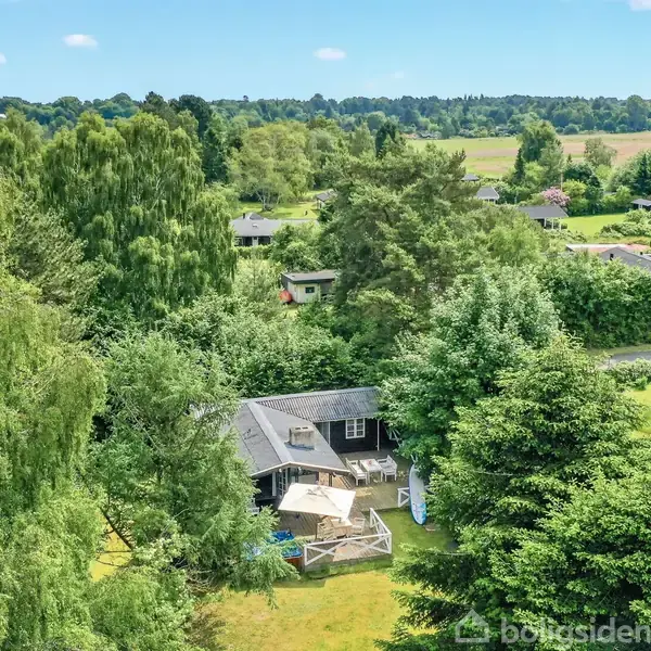Sort sommerhus med åben dør til træterrasse, hvor der står en parasol over et bord og stole. En jacuzzi er placeret til venstre, og en surfbræt står op ad hegnet.