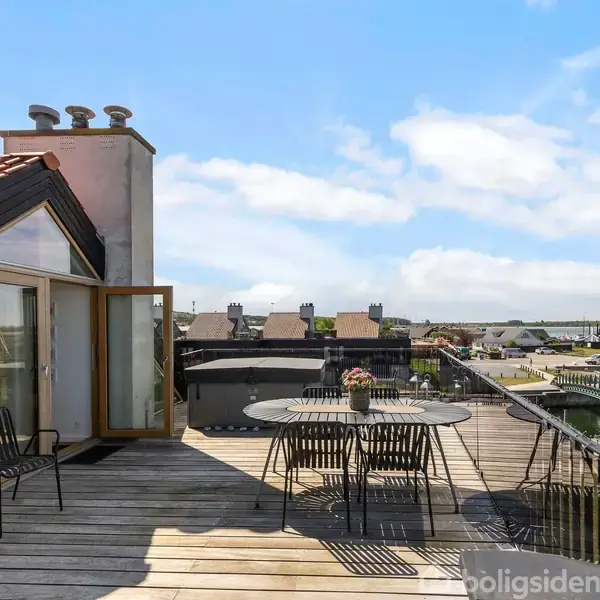En møbleret tagterrasse med bord og stole står under en blå himmel med skyer, omgivet af en marina med både og huse i baggrunden.