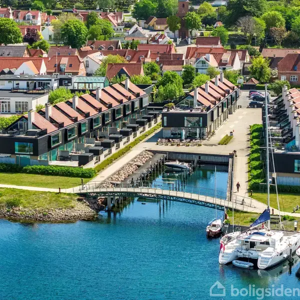 Rækkehuse ligger langs en smal kanal, omgivet af grønne områder og parkerede biler. Foran er en lille bro, flere både ligger ved kaj langs en bugt. Bagved ses en by med røde tage.