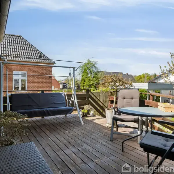 Terrasse med træplanker har en gyngesofa, et rundt bord, og stole. Omkranset af planter og have, med udsigt til et murstenshus og træer under en blå himmel.