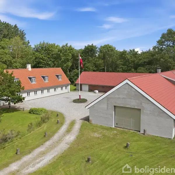 Et hvidt landhus med rødt tag står ved siden af en stor lade med rødt tag. En dansk flagstang står i en gårdsplads omgivet af grønne træer og græsplæne.