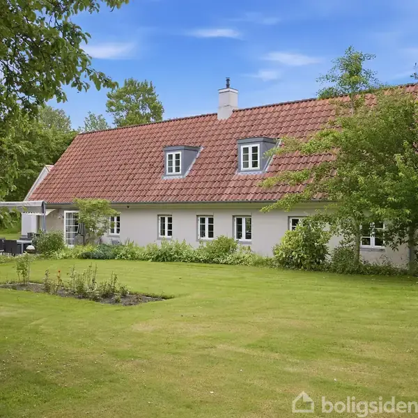 Hvidt hus med rødt tegltag står stille omgivet af en grøn have. Træer og buske omkranser byggeriet, og en terrasse ses til venstre. Himmel er blå.