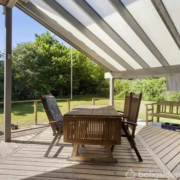 Terrasse med træbord og stole under overdække, omgivet af grønne træer og græsplæne. I baggrunden står en bænk. Sollys skinner gennem det gennemsigtige tag. Tekst: Boligsiden.