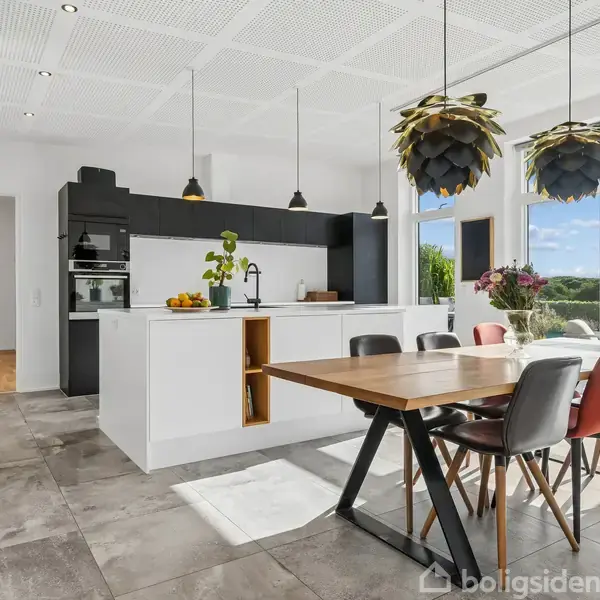 Et moderne køkken med en stor ø og spisebord; hængelamper over bordet. Store vinduer vender ud mod en have og blå himmel. Indretningen er lys og minimalistisk.