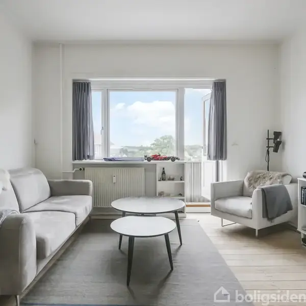 Sofa og stol står i stue, rettet mod et stort vindue. TV og reol til højre, bord i midten. Lysegråt tæppe på gulvet, bøger i reolen.