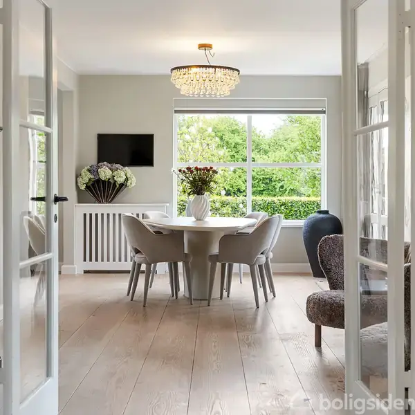 Spisebord med fem stole omkranser en vase med blomster. Rummet har lyse vægge, en lysekrone, og et stort vindue med udsigt til grønt. En væg dekoreret med en fladskærm.
