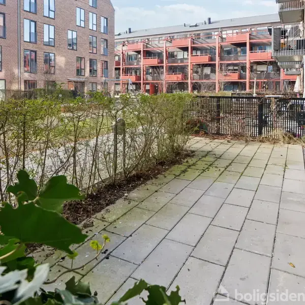 En terrassesessionser område med fliser og lav hæk. I baggrunden ses moderne murstensbygninger med balkoner, omgivet af et fælles græsareal.