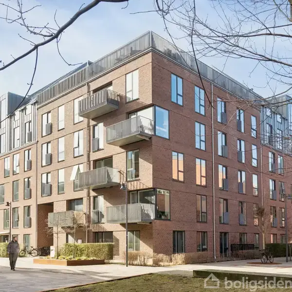 En moderne boligbygning med store vinduer og balkon. En person går på fortovet foran bygningen. Træer uden blade omgiver området, der er lyst og bymæssigt.
