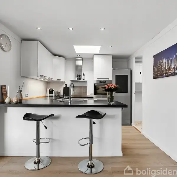 Et moderne køkken med to barstole ved en sort køkkenø. I baggrunden ses hvide skabe og en indbygget ovn. Til højre hænger et bybillede på væggen.