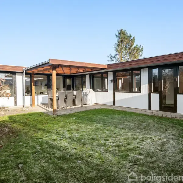 En moderne etplansvilla med store vinduer omkranser en grønt haveområde. En overdækket terrasse har spisebord og stole. Himlen er blå, hvilket indikerer en solrig dag.