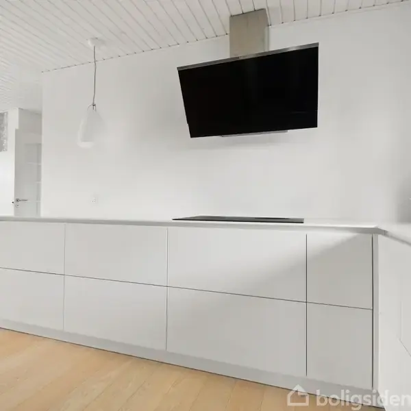 TV hænger på en hvid væg i et moderne køkken. Under TV'et er en minimalistisk køkkenbordplade med grå skuffer. Lyse trægulve og en hængende lampe ses i baggrunden.