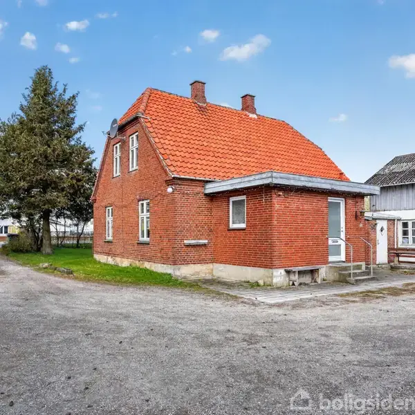 Murstenshus med rødt tag står ved grusvej. Huset har flere vinduer, og en tilhørende terrasse er synlig. Omgivelserne inkluderer træer og andre bygninger.