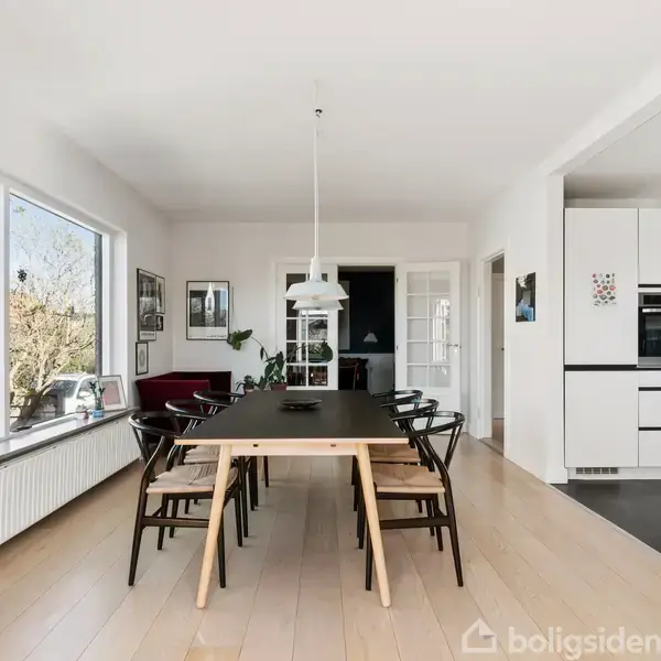 Et langt spisebord med sorte stole står i midten af et lyst, moderne køkken-alrum med store vinduer og trægulv. En lampe hænger over bordet, omgivet af køkkenudstyr og dekorationer.