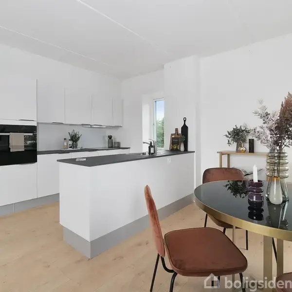 Et moderne køkken har hvide skabe og en central ø med grå bordplade. En rund spisebord med brune stole står i forgrunden. Flasker og planter pynter rummet.