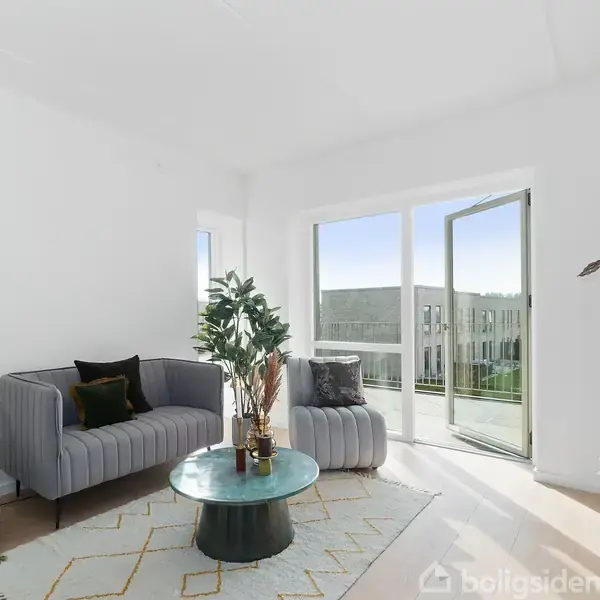 En stue med en grå sofa og et lille sofabord med planter. Rummet er lyst med åbne glasdøre til en altan og udsigt til moderne bygninger. En reol står ved siden af.