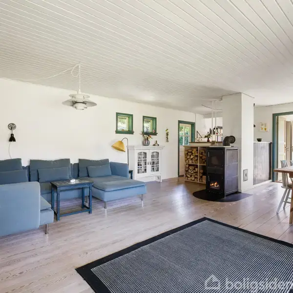 Stue med blå sofa og sort pejseovn. Spisebord med stole til højre. Hvide vægge og trægulv skaber et lyst og åbent rum. Store vinduer giver udsigt til have.