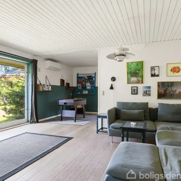 En stue med grå sofa og vægkunst, står ved siden af et stort vindue, der viser ud til en grøn have. En bordfodboldspil står i hjørnet, og der er trægulve.