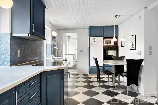 Et moderne køkken med mørkeblå skabe og skuffer, udfører en funktionel og stilfuld atmosfære. En rund spisebord med sorte stole er placeret ved siden af væggen med ovn og skabsplads. Skakternet gulv og hvidt loft tilføjer kontrast.