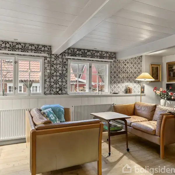En stue med to lædersofaer og et sofabord. Rummet er lyst med vinduer, blomstrede tapeter og vægmalerier. En lampe står på bordet ved siden af sofaen, og en blå skål på et sidebord.