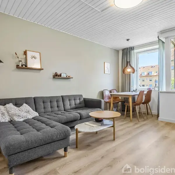 Grå hjørnesofa med puder står i en stue. Foran er et rundt sofabord. Ved siden af spiseskab med brune stole og lyse pendellamper. Store vinduer åbner mod altan.