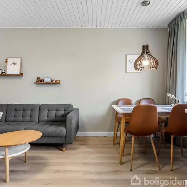 Grå sofa står i stuehjørnet med to små borde foran. En spisebord med fire brune stole ved siden af. Hylder med dekorationer hænger på væggen. Store vinduer giver lys.