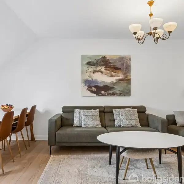 En stue med to grå sofaer prydet med puder, er centralt placeret. På væggen hænger et maleri, og en lysekrone hænger over et sofabord i en rummelig, lys spiseområde.