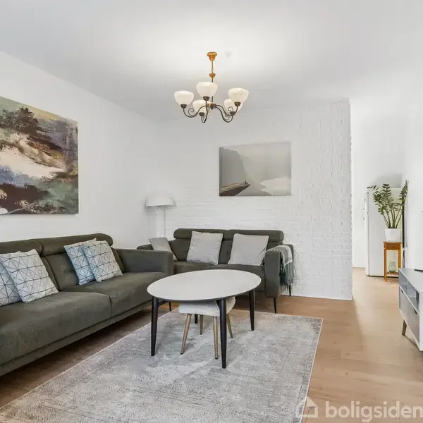 Sofa står i stue, omgivet af sofabord og tæppe. Væggene er dekoreret med malerier, og en standerlampe står i hjørnet. Et TV er placeret på møbel.