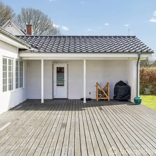 Terrasse af træplanker foran et hvidt hus med store vinduer, en dør i midten. En grill dækket med sort betræk står i hjørnet. Græsplæne og hæk i baggrunden.