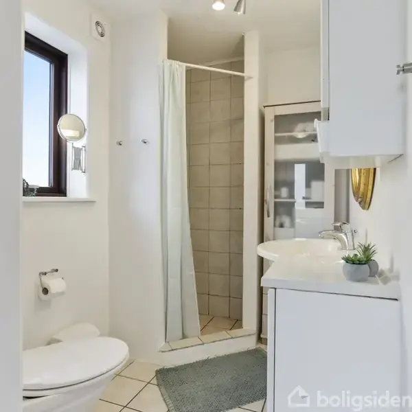 Toilet mod væg, bruser med forhæng i hjørnet; ved siden af er der et håndvaskeskab med spejl. Lyse fliser og et vindue til venstre. Ingen tekst til stede.