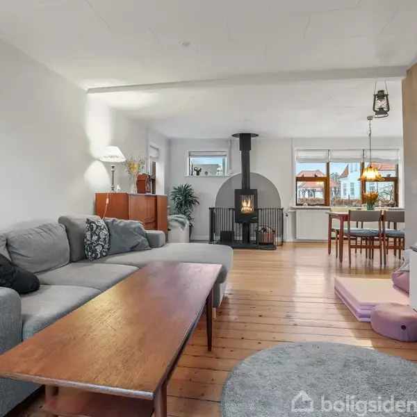 En grå sofa står ved et træbord i en stue, hvor en brændeovn opvarmer rummet med spiseplads bagved. Store vinduer giver naturligt lys, og træmøbler fremhæver interiøret.