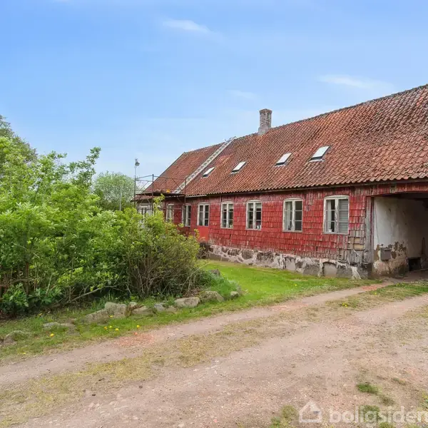 Rødt murstenshus med skråtag, omgivet af grusvej og buske, står i landlig, åbent område med grønne træer i baggrunden.