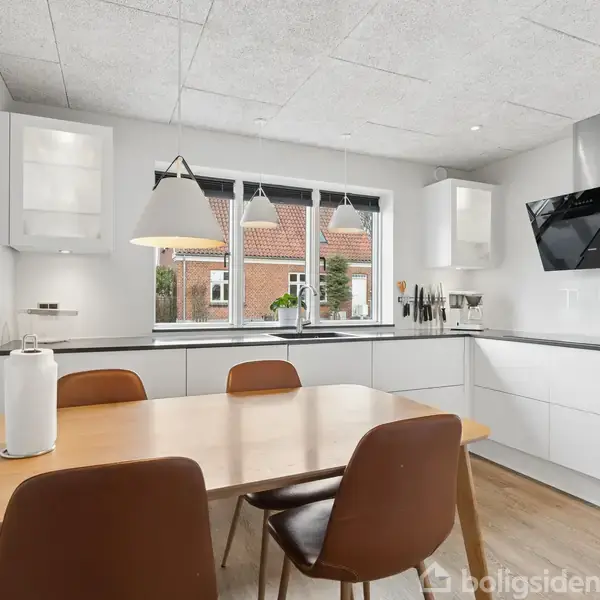 Et spisebord med en blå dug står centralt i et lyst rum. Seks stole omgiver bordet, og en moderne lampe hænger over det. Maleri og altan ses i baggrunden.