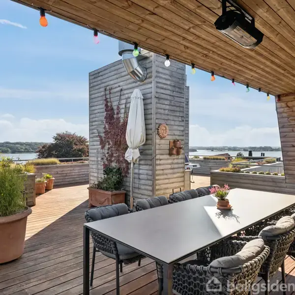 En udendørs terrasse med et stort spisebord omgivet af stole. Overdækket af et træloft med farverige lyskæder. Omkring omgivelserne ses blomsterkrukker og udsigt over en sø.