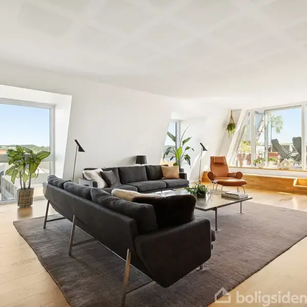 Sofa i sort læder står på et tæppe i en lys stue med trægulv, omgivet af planter og glasdøre, der åbner til en balkon med udsigt.