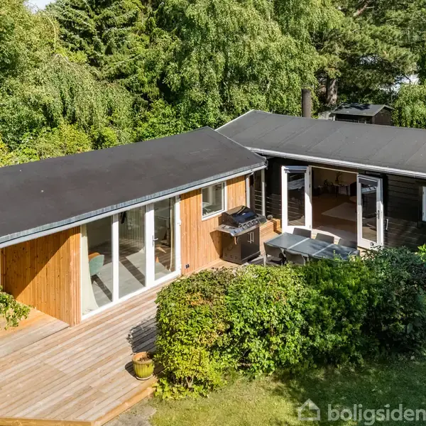 Et moderne træhus med fladt tag har en stor træterrasse med grill og havemøbler. Det er omgivet af tæt, grøn beplantning og træer i en villaagtig have.