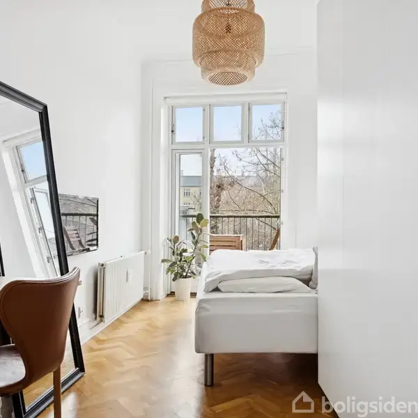 Et lyst soveværelse med en seng til højre, et skrivebord med stol til venstre og et stort spejl stående op ad væggen. Et vindue i baggrunden giver udsigt til træer.