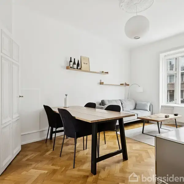 Et lyst spise- og opholdsområde med et spisebord, fire stole, en grå sofa og et sofabord. Hylder med dekoration hænger på en hvid væg, og store vinduer lukker naturligt lys ind.