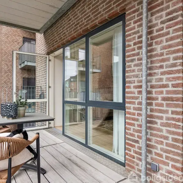 Altan med et bord og stole i flettet design. Det åbne glasparti leder til en lejlighed. Omgivet af murstensbygning med flere altaner i et tæt boligkompleks.