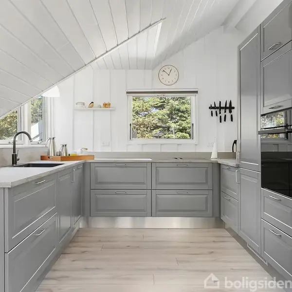Et moderne køkken med grå skabe og hvide vægge. En stor vask, komfur og vægur. Vinduerne viser en udsigt til træer udenfor. Ingen tekst er fundet på skabene.
